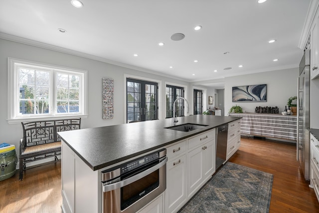 kitchen with stainless steel appliances, dark countertops, white cabinetry, a sink, and a kitchen island with sink