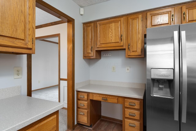 kitchen featuring built in desk, stainless steel fridge with ice dispenser, light countertops, brown cabinetry, and baseboards