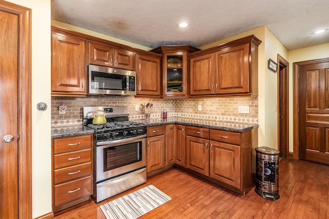 kitchen with light wood-style floors, appliances with stainless steel finishes, brown cabinets, dark stone counters, and glass insert cabinets