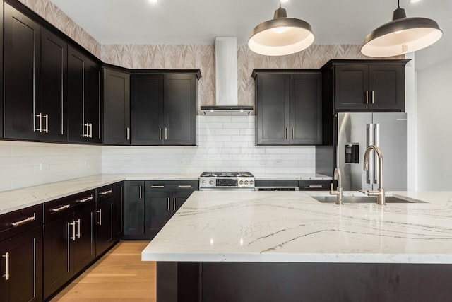 kitchen featuring decorative light fixtures, appliances with stainless steel finishes, a sink, wall chimney range hood, and light stone countertops