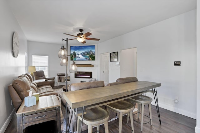 dining space with a ceiling fan, a glass covered fireplace, dark wood-style flooring, and baseboards