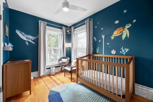 bedroom featuring hardwood / wood-style floors, a nursery area, a ceiling fan, and baseboards