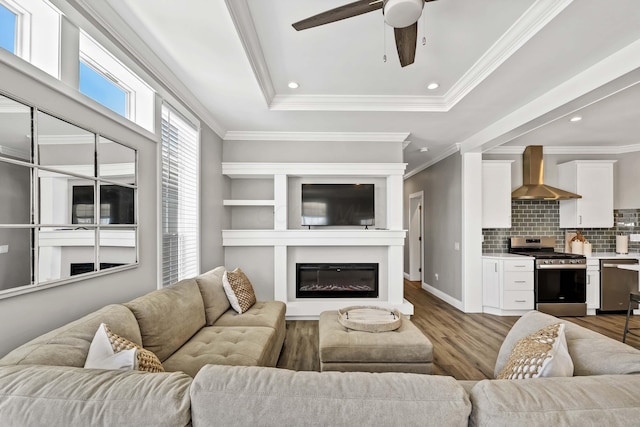 living room featuring a tray ceiling, light wood finished floors, ornamental molding, a glass covered fireplace, and ceiling fan