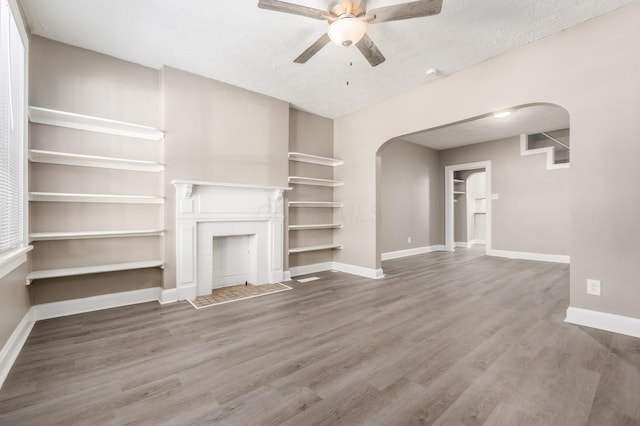 unfurnished living room featuring ceiling fan, baseboards, a fireplace with flush hearth, wood finished floors, and arched walkways