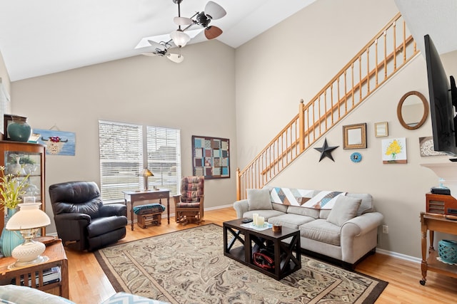 living area featuring baseboards, ceiling fan, light wood-style flooring, stairs, and high vaulted ceiling