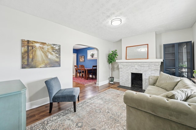 living room featuring arched walkways, a textured ceiling, wood finished floors, baseboards, and a brick fireplace