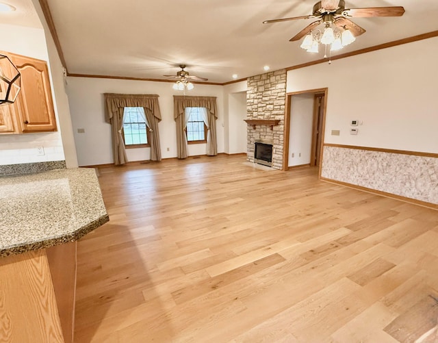 unfurnished living room with ornamental molding, a stone fireplace, wainscoting, light wood-style flooring, and a ceiling fan