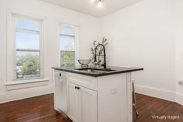 kitchen with white dishwasher, a kitchen island with sink, a sink, white cabinets, and dark countertops