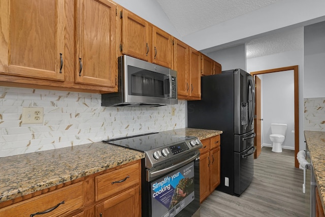 kitchen with a textured ceiling, light wood-style flooring, appliances with stainless steel finishes, brown cabinets, and dark stone counters