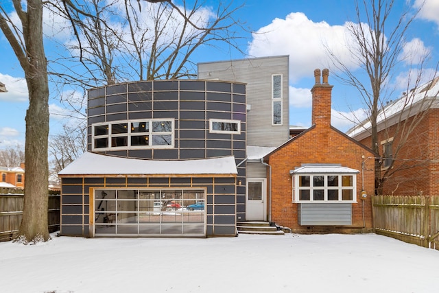 snow covered rear of property with a garage, brick siding, a chimney, and fence