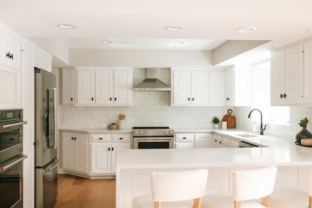 kitchen featuring appliances with stainless steel finishes, a breakfast bar area, a peninsula, light countertops, and wall chimney range hood