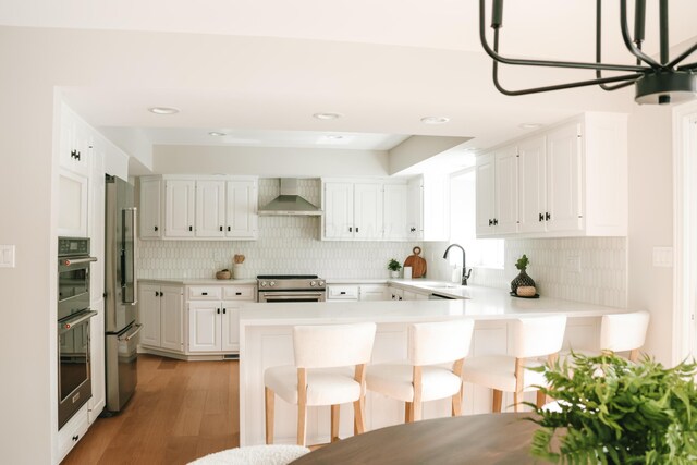 kitchen featuring a peninsula, stainless steel appliances, wall chimney range hood, a kitchen bar, and a sink