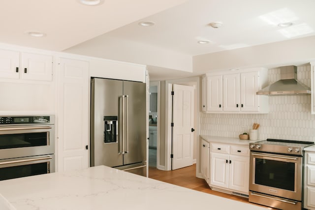 kitchen featuring tasteful backsplash, white cabinets, wall chimney exhaust hood, appliances with stainless steel finishes, and light stone countertops