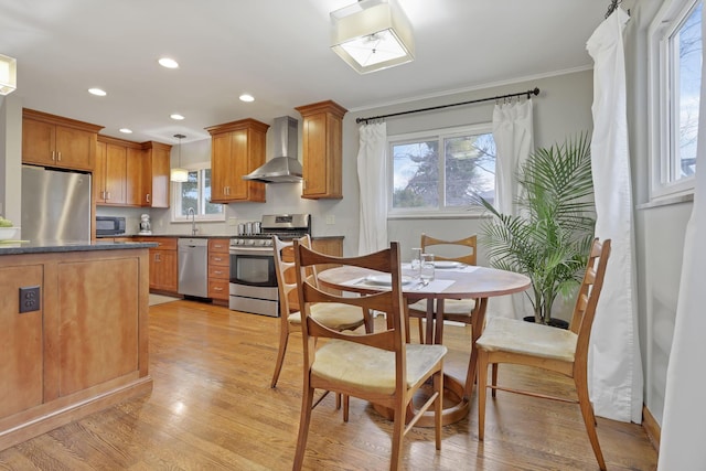 kitchen with stainless steel appliances, wall chimney range hood, brown cabinets, light wood finished floors, and dark countertops