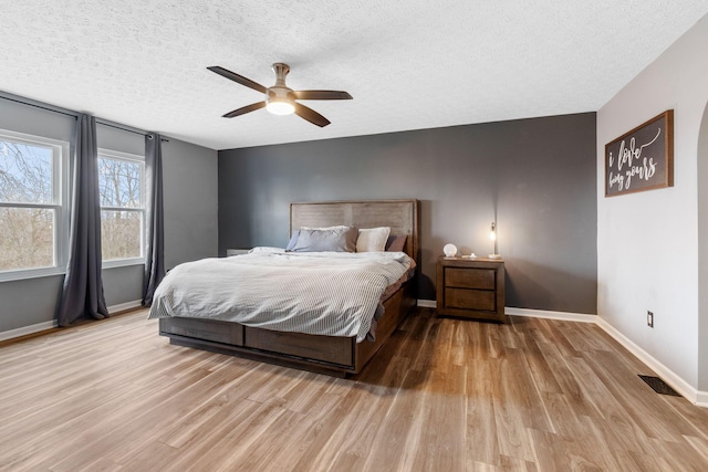 bedroom with a textured ceiling, light wood-style flooring, a ceiling fan, visible vents, and baseboards