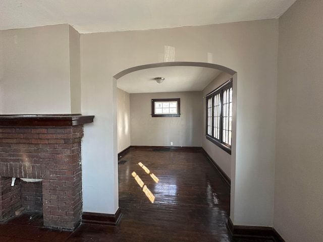 unfurnished living room with dark wood-style floors, a brick fireplace, baseboards, and arched walkways