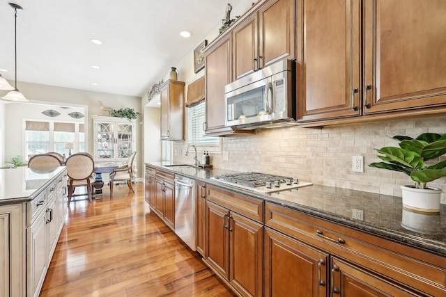 kitchen featuring brown cabinetry, dark stone countertops, stainless steel appliances, light wood-style floors, and pendant lighting