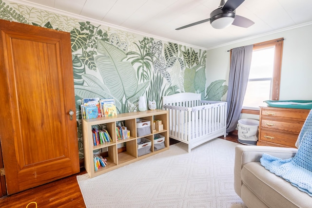 bedroom featuring crown molding, ceiling fan, wood finished floors, a nursery area, and wallpapered walls