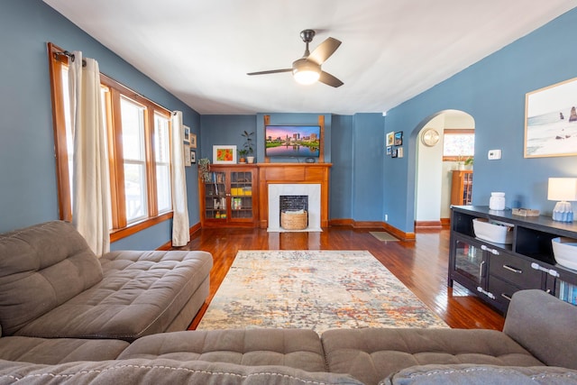 living area featuring arched walkways, wood finished floors, a fireplace with flush hearth, a ceiling fan, and baseboards