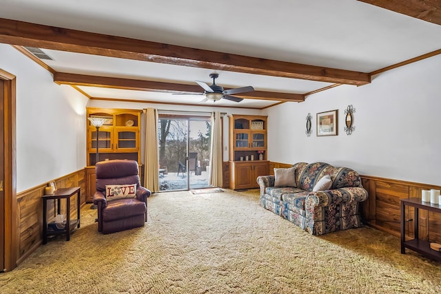 carpeted living area with ornamental molding, a ceiling fan, wainscoting, wood walls, and beamed ceiling