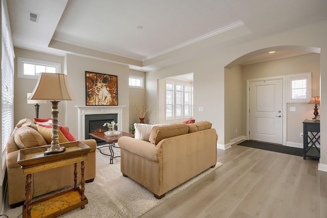 living area with arched walkways, a raised ceiling, light wood-style flooring, a lit fireplace, and baseboards