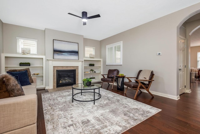 living room with dark wood-style floors, arched walkways, ceiling fan, a tile fireplace, and baseboards