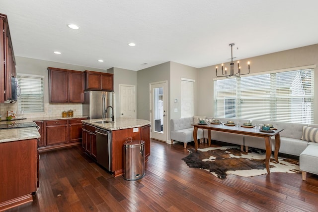 kitchen with appliances with stainless steel finishes, open floor plan, light stone counters, hanging light fixtures, and a kitchen island with sink