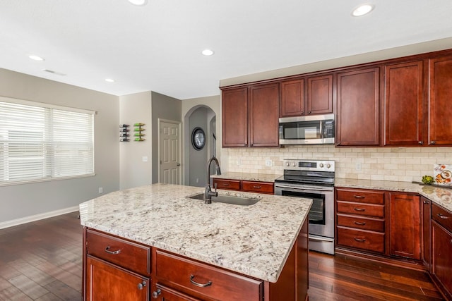 kitchen with stainless steel appliances, dark wood-type flooring, a sink, an island with sink, and light stone countertops