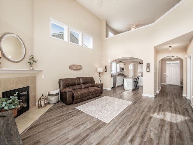 living area with baseboards, arched walkways, a tile fireplace, a towering ceiling, and wood finished floors