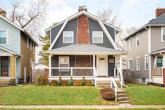 dutch colonial with a gambrel roof, a chimney, roof with shingles, covered porch, and a front yard