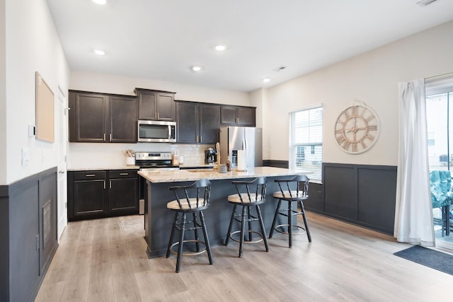 kitchen with light stone counters, a breakfast bar, appliances with stainless steel finishes, light wood-style floors, and an island with sink