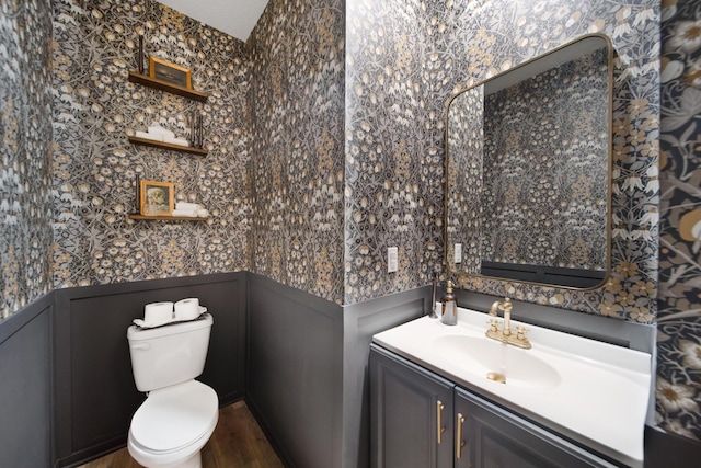 half bath with toilet, wallpapered walls, a wainscoted wall, and vanity