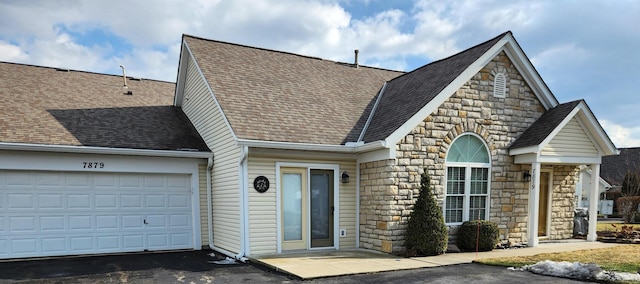 view of front facade featuring stone siding, roof with shingles, an attached garage, and driveway