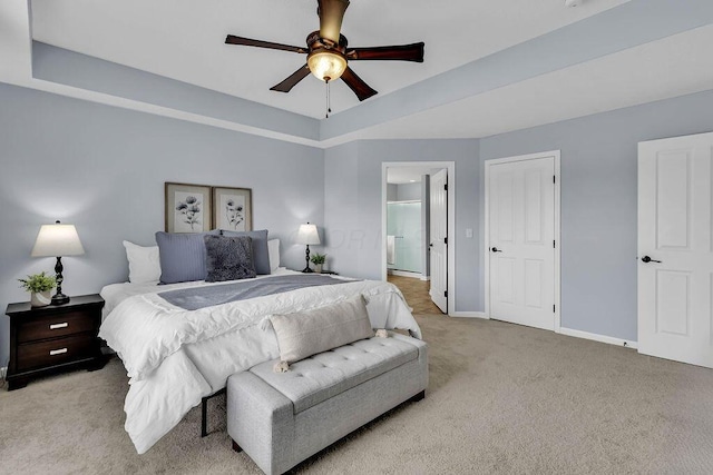 bedroom with ceiling fan, light colored carpet, baseboards, a raised ceiling, and ensuite bath