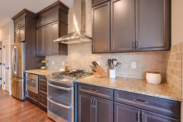 kitchen featuring decorative backsplash, wall chimney exhaust hood, appliances with stainless steel finishes, wood finished floors, and light stone countertops