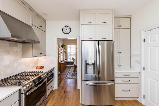 kitchen with light countertops, appliances with stainless steel finishes, light wood-type flooring, wall chimney exhaust hood, and tasteful backsplash