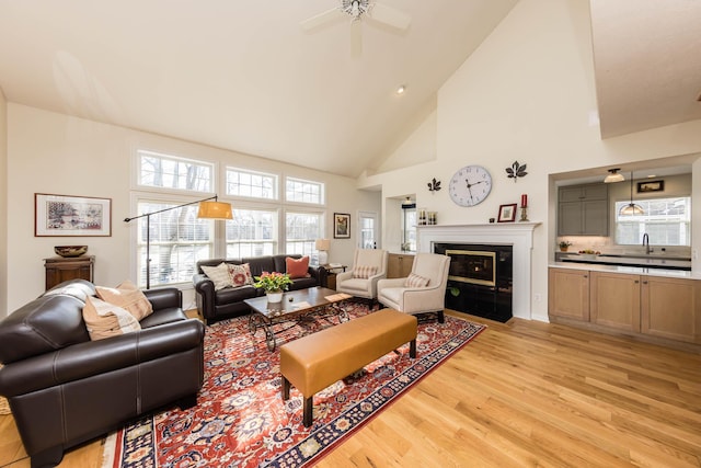 living area with ceiling fan, high vaulted ceiling, a fireplace with flush hearth, and light wood-style flooring