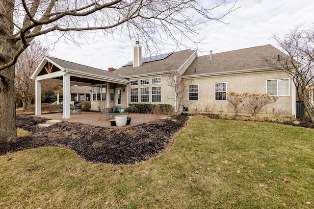 back of house featuring a chimney, a yard, a patio area, roof mounted solar panels, and stucco siding