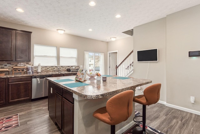 kitchen featuring dark brown cabinetry, wood finished floors, backsplash, dishwasher, and a kitchen bar