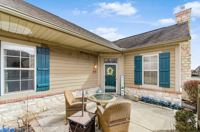 view of exterior entry with a shingled roof, a chimney, a patio area, and brick siding