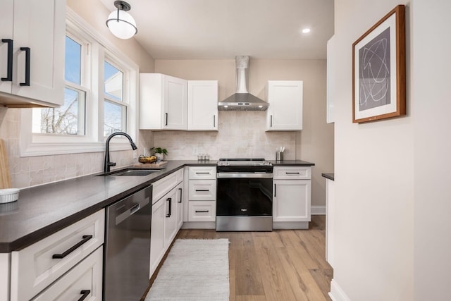 kitchen featuring dark countertops, appliances with stainless steel finishes, white cabinetry, a sink, and wall chimney range hood