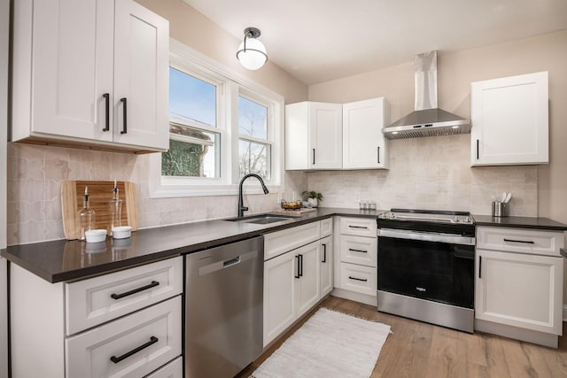 kitchen with a sink, appliances with stainless steel finishes, light wood-type flooring, wall chimney exhaust hood, and dark countertops