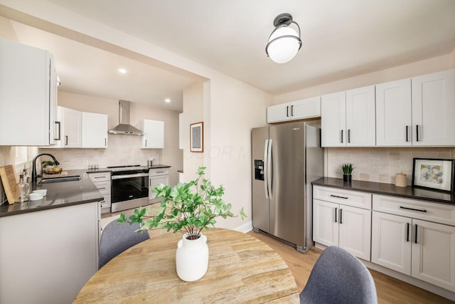 kitchen with dark countertops, appliances with stainless steel finishes, white cabinets, a sink, and wall chimney range hood