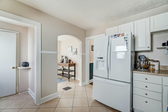 kitchen featuring arched walkways, white refrigerator with ice dispenser, light tile patterned floors, white cabinetry, and baseboards