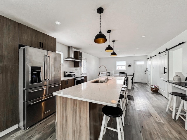 kitchen featuring a barn door, appliances with stainless steel finishes, wall chimney range hood, a kitchen bar, and a sink