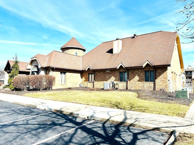 view of front facade featuring brick siding, roof with shingles, uncovered parking, a chimney, and a front yard