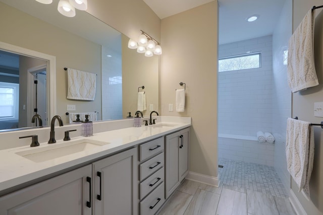 full bathroom with a tile shower, double vanity, a sink, and baseboards