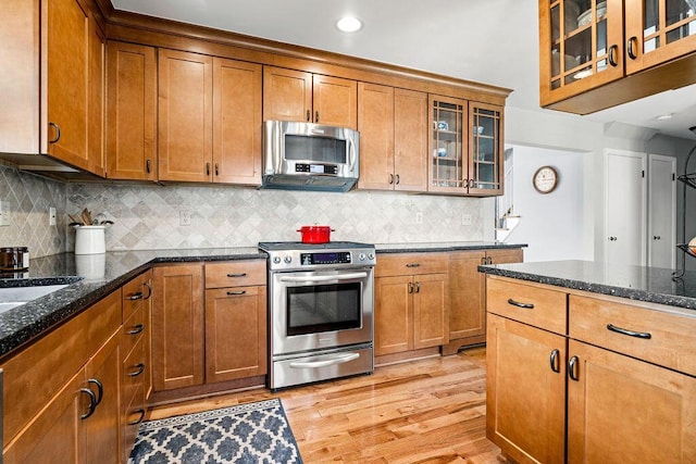 kitchen with appliances with stainless steel finishes, light wood-type flooring, backsplash, brown cabinets, and dark stone countertops