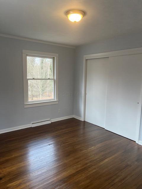 unfurnished bedroom with a baseboard heating unit, dark wood finished floors, a closet, and ornamental molding