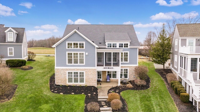 rear view of house with a sunroom, stone siding, roof with shingles, a lawn, and a patio area